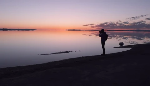 Silhouetted photographer standing on a beach at dusk, facing a calm mirror-like water at twilight.