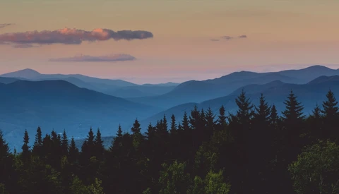 Layered mountain ridges receding into blue-purple haze at sunset, with dark coniferous forest silhouetted in foreground.
