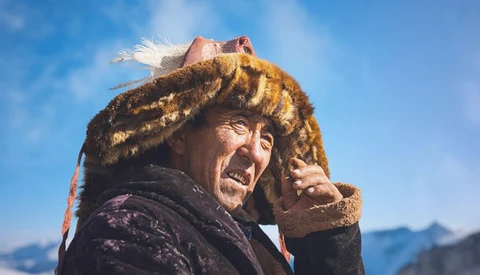 Person wearing a fur hat with animal ears smiles at camera against blue sky and snowy mountains.
