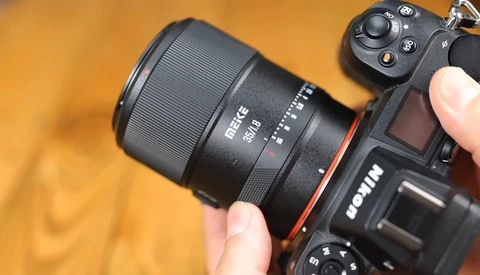 Prime lens mounted on mirrorless camera body held in hands against warm yellow background.