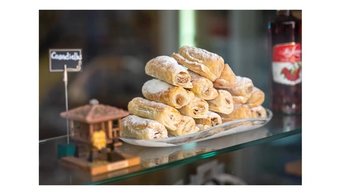 Stacked pastry rolls dusted with powdered sugar displayed on a white plate behind a glass bakery counter.