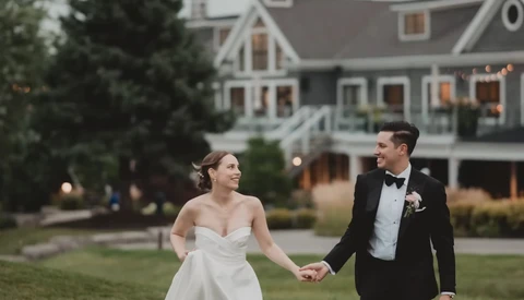 Bride and groom holding hands while walking on manicured lawn in front of large residential home at dusk.
