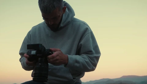 Photographer holding a professional video camera during golden hour with mountains visible in the background.