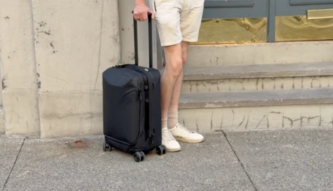 Person standing with black rolling carry-on luggage on concrete pavement beside a building.