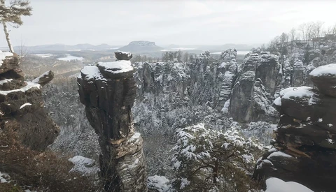 Snow-dusted sandstone rock formations rise from a forested valley under overcast winter sky.