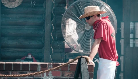 Man in straw hat and red shirt leaning against weathered teal wooden structure with large industrial fan.