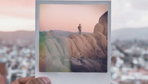 Polaroid photograph held in front of a blurred cityscape, showing a lone figure standing atop a large rock formation at sunset.