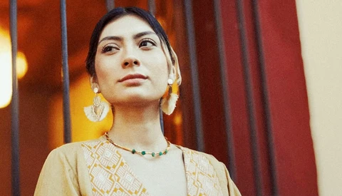 Woman wearing a patterned beige tunic and statement earrings gazes upward against warm amber and red draped backdrop.