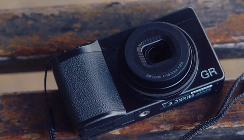 Ricoh GR compact camera with leather grip and neck strap on weathered wooden surface.