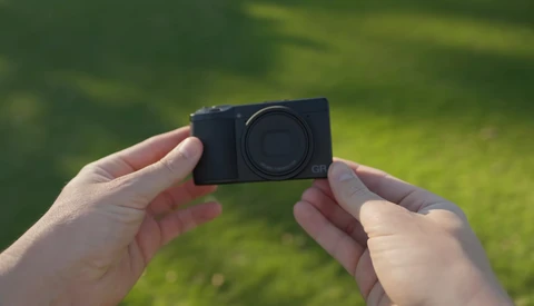 Hands holding a compact black mirrorless camera body against a blurred green outdoor background.