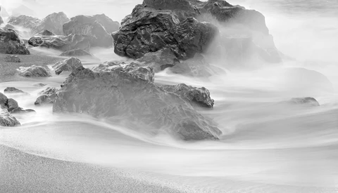 Rocky coastal rocks shrouded in soft, flowing ocean mist and waves on a sandy beach.