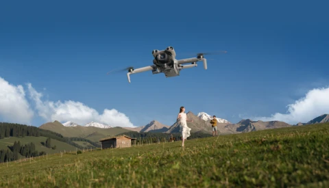Drone flying above a grassy alpine meadow with mountains in the background and people standing on the ground.