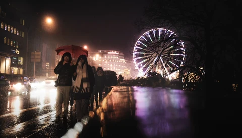 Wet city street at night with illuminated Ferris wheel reflecting in water and pedestrians in foreground.