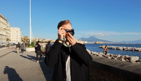 Person in black blazer and sunglasses holding camera to face on waterfront promenade.