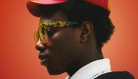 Profile portrait of a person wearing yellow-tinted sunglasses, red cap, and white collar against warm orange background.