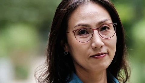 Woman wearing rose-gold rimmed glasses and blue shirt, smiling slightly at camera with soft natural outdoor lighting.