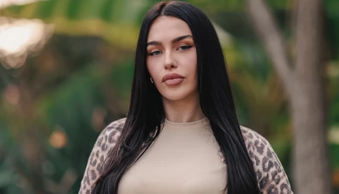 Woman with long black hair wearing a cream-colored top and leopard-print cardigan, photographed outdoors with blurred green foliage background.