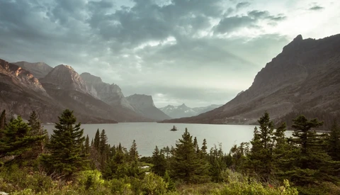 Mountain lake surrounded by evergreen forest with dramatic peaks and storm clouds overhead.