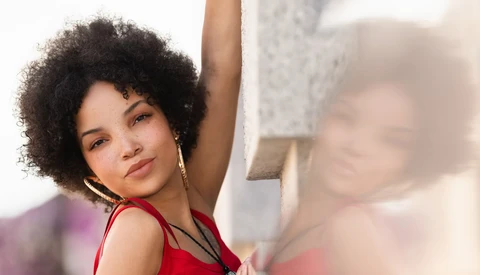 Woman with natural curly hair wearing red sleeveless top, arm raised, looking at camera with soft expression.