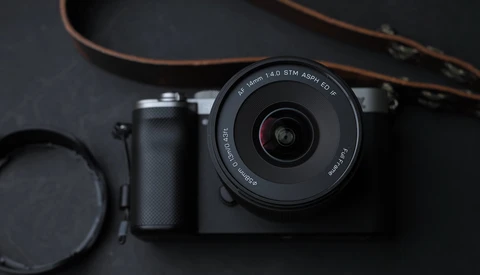 Mirrorless camera with prime lens, lens cap, and brown leather strap on dark surface.