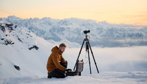 Photographer in brown jacket sitting in snow with camera mounted on tripod, mountain range and cloud inversion visible in background.