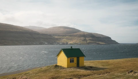 Bright yellow house with green roof on barren coastline beside dark ocean and distant mountains.