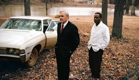 Two men in business attire standing beside a white sedan in a wooded area with a pond visible in the background.