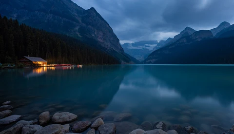 Mountain lake at twilight with illuminated boathouse reflected in perfectly still turquoise water.