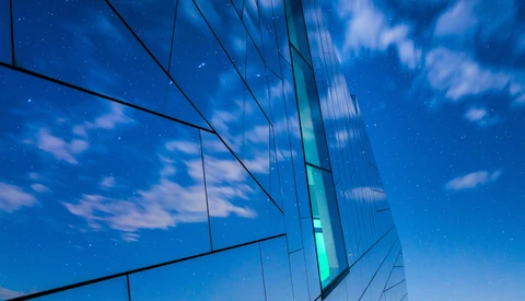 Modern glass building facade reflecting blue sky and clouds with geometric lines and reflections.