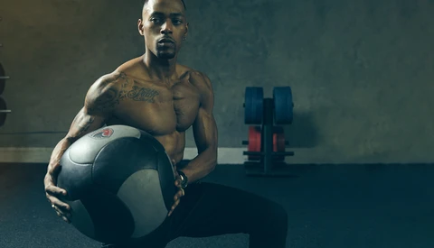Shirtless man holding a medicine ball in a gym with dumbbells visible in the background.