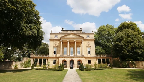 Neoclassical stone mansion with columned portico, symmetrical facade, and manicured grounds under blue sky.