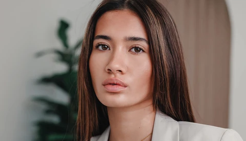 Woman with long dark hair wearing a white blazer, gazing directly at camera with neutral expression.