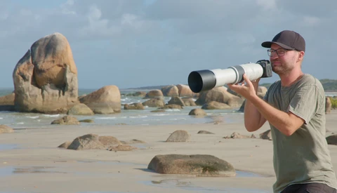 Photographer holding a telephoto lens on a beach with large granite rock formations.