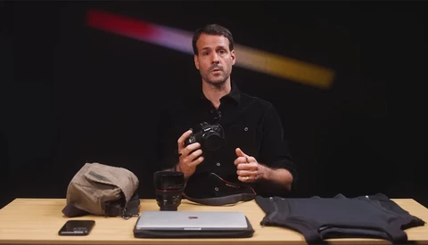 Man in black shirt gesturing while speaking at table with photography gear and laptop.