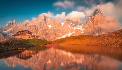 Alpine peaks with snow reflected in a still lake at golden hour, with a wooden lodge on the shoreline.
