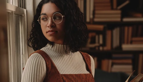 Portrait of a person wearing round glasses and a cream ribbed turtleneck with a rust-colored vest, positioned against bookshelves.