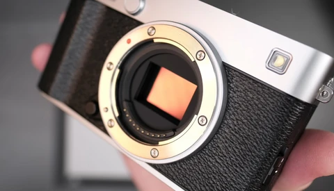 Mirrorless camera with exposed sensor and open body cap, held in hands against neutral background.