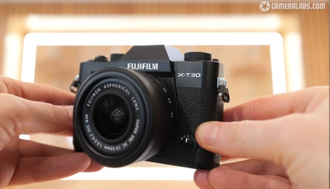 Hands holding a Fujifilm X-T30 mirrorless camera with attached lens against a blurred warm-toned background.