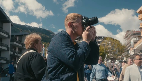 Photographer with camera raised to eye, focusing on subject during outdoor event with crowd and mountains in background.