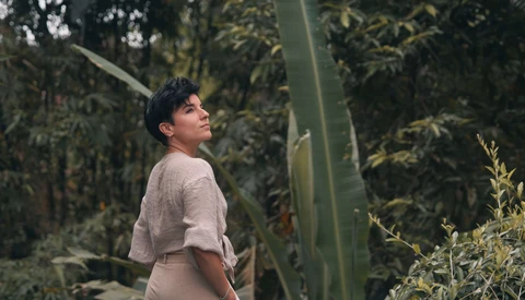 Person in beige shirt standing among tall green plants and cacti in a greenhouse environment.