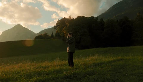 Person standing in a grassy meadow with forested mountains in the background during golden hour.