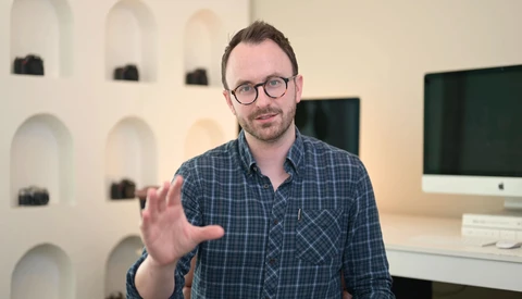 Man wearing glasses and plaid shirt gestures while speaking in a studio setting.