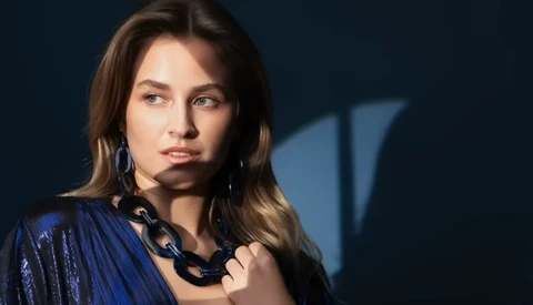 Woman with long brown hair wearing a blue sequined jacket and black chain necklace against a dark blue background.