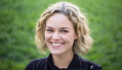 Young woman with shoulder-length wavy blonde hair smiling at camera against blurred green background.