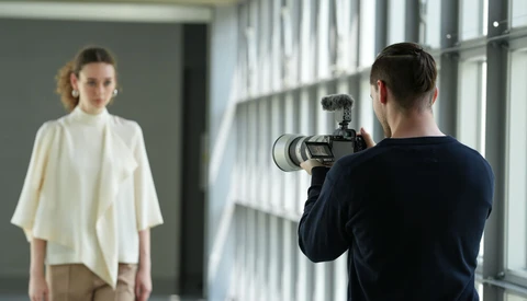 Photographer operating a camera on a shoulder rig while capturing a portrait of a woman in a bright, minimalist studio.