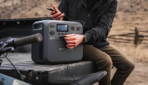Person holding a portable power station with digital display while sitting in truck bed outdoors.