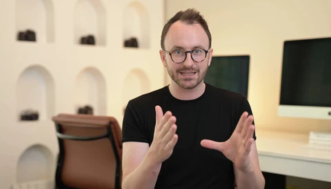 Man in black t-shirt and glasses gesturing while speaking in a modern studio setting.