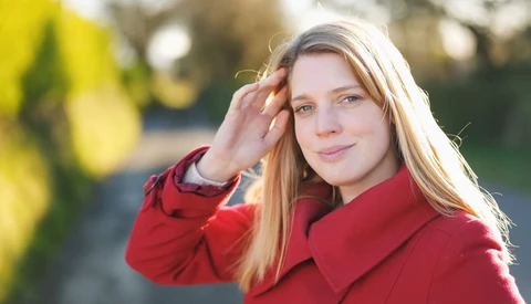 Woman with long strawberry blonde hair wearing a red coat, touching her hair and looking at camera outdoors.
