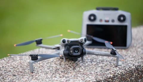 Compact folding drone with camera positioned on gravel surface, remote controller blurred in background against green foliage.