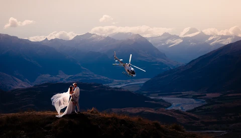 Bride and groom standing on a mountain ridge with a helicopter flying nearby and snow-capped peaks in the distance.